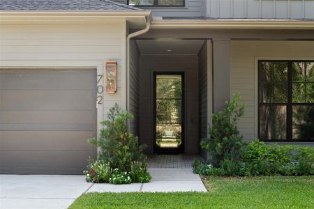 This home features a modern exterior with a neutral color palette, a large garage door, and an inviting entryway framed by lush landscaping. The window provides a glimpse of the greenery outside, enhancing the home's connection with nature.