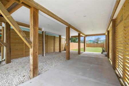 This is a covered carport area with a concrete floor, featuring wooden support beams and a wooden privacy fence, leading into a spacious backyard with a, green lawn.