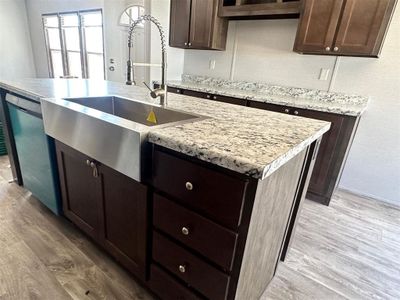 Kitchen with dishwasher, a kitchen island with sink, light stone countertops, dark brown cabinetry, and light wood-type flooring