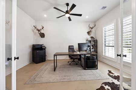 The modern and well-lit home office featuring a large window with plantation shutters, a ceiling fan, and optimal space for desk setup.