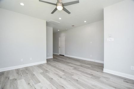 This is a spacious, main living room featuring light gray walls, contemporary wood-look flooring, and a ceiling fan with lighting. There's ample natural light and a doorway leading to another part of the home. The room is a blank canvas ready for personalization.