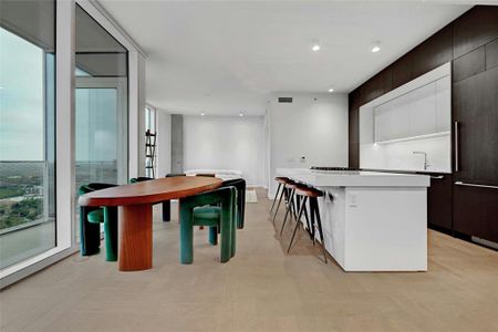 A modern, open-concept kitchen, dining and living area. The kitchen features a sleek, minimalist design with a large island at its center, topped with white-colored stone countertop. The island includes a built-in gas cooktop and stainless steel oven. *Linden Residence No. 2406 Furnished Model Shown Here (Furnishings provided by Scott + Cooner / Cassina Available for Purchase)