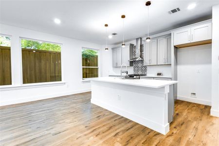 Kitchen featuring wall chimney range hood, light hardwood / wood-style flooring, an island with sink, decorative light fixtures, and backsplash