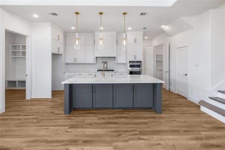 Kitchen with light wood-type flooring, an island with sink, stainless steel double oven, white cabinetry, and hanging light fixtures