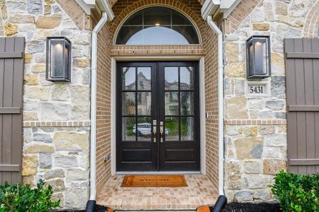 This is the front entrance of the home featuring a stately double door with glass panes, flanked by stone and brickwork, and complemented by elegant outdoor sconces. The welcoming atmosphere is enhanced by an arched transom window above the door. The house number is prominently displayed.