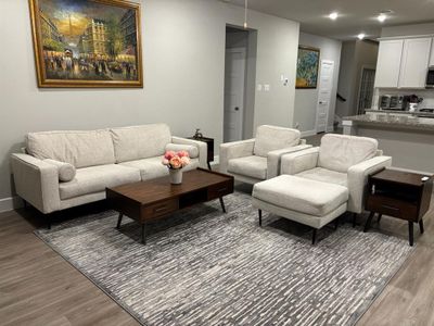 This is a modern living room featuring a neutral color palette. Showcasing an open-concept layout that flows into a well-lit kitchen with white cabinetry.