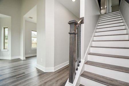 This is a modern interior space featuring a staircase with dark wood accents and metal balusters, complemented by light gray walls and laminate wood flooring. The area is well-lit with natural light from windows and recessed ceiling lights.