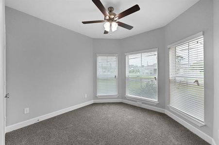 This is a bright, empty room with neutral gray walls, carpeted flooring, three large windows with blinds, and a ceiling fan with lighting. It appears clean and ready for move-in.