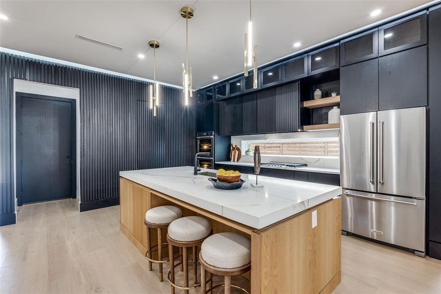 Kitchen featuring light stone countertops, light hardwood / wood-style flooring, stainless steel fridge, a center island with sink, and pendant lighting