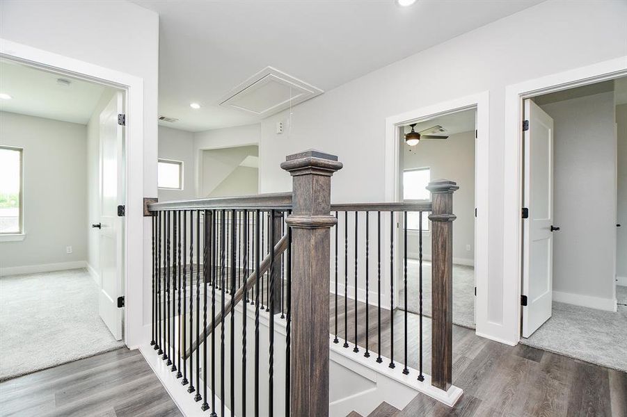 This photo showcases an upper-level interior space with a neutral color palette, featuring an open landing with dark-stained wooden railing, plush carpeting in the adjacent room, and modern flooring in the hallway. Multiple doors suggest access to several rooms, and the space is well-lit with natural and artificial light.