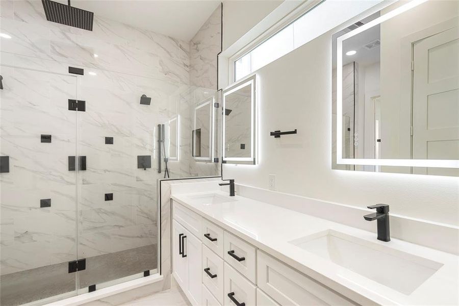 This is a modern, sleek bathroom featuring marble-like tiled walls, a glass-enclosed shower, double vanity with white cabinets, black fixtures, and backlit mirrors.