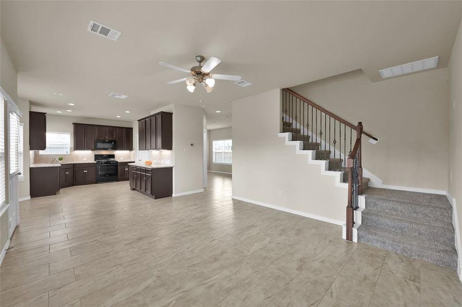 This is a spacious, open-concept interior featuring a modern kitchen with dark cabinetry and, tile flooring throughout, and a staircase leading to the upper level. The area is well-lit with natural light from windows and includes a ceiling fan.