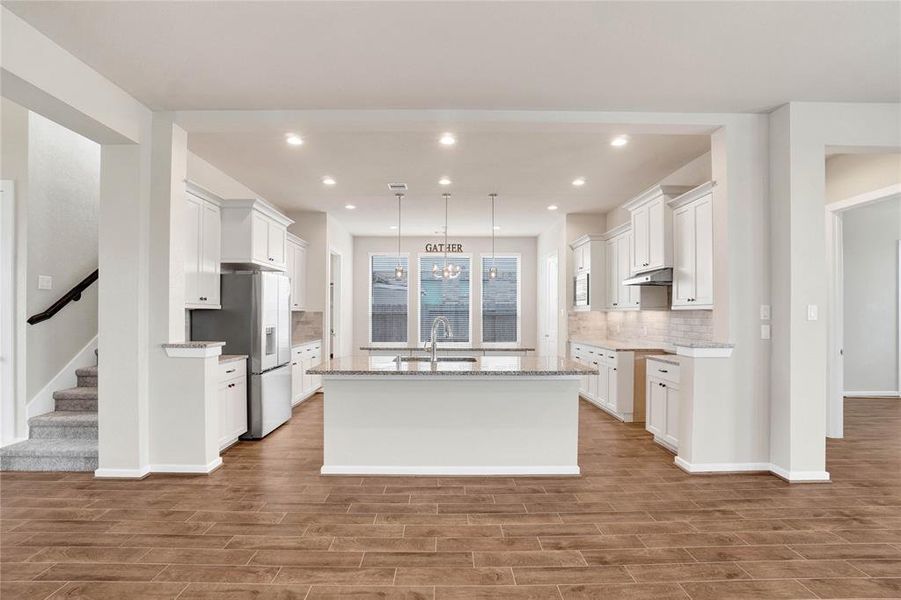 This is a spacious, modern kitchen with white cabinetry, granite countertops, and stainless steel appliances. It features a large island with pendant lighting and opens up to a well-lit dining area with windows. The room has wood flooring and a neutral color palette, ideal for personal customization.