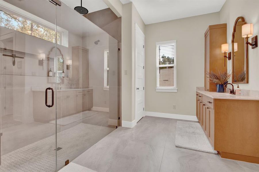 The primary bathroom features a large walk-in shower enclosed by glass doors, identical vanities, warm wood cabinetry, and stylish fixtures. The space is well-lit and has a neutral color palette.