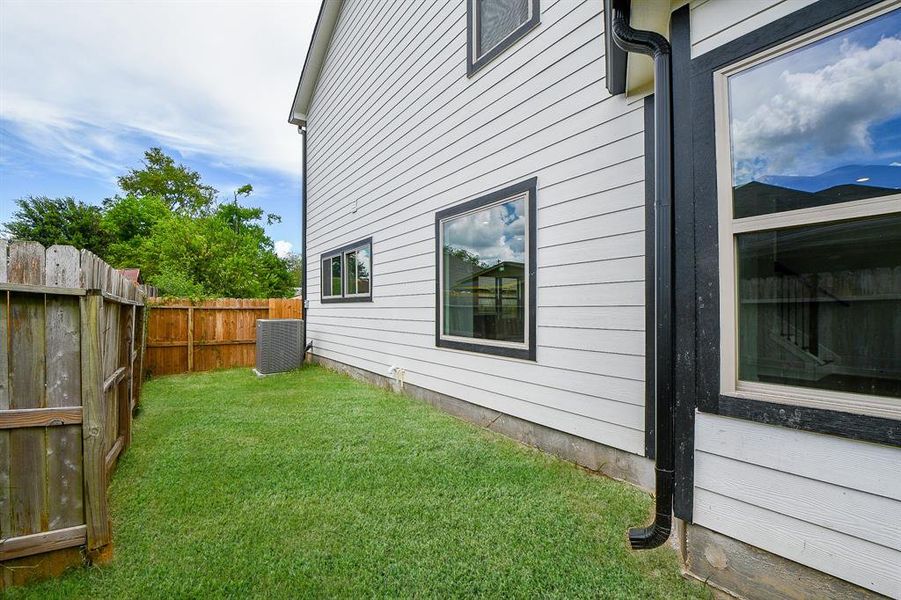 This is a well-maintained side yard with a lush green lawn, bordered by a wooden fence for privacy. The exterior of the home features clean, modern siding with multiple windows providing natural light inside. An air conditioning unit is present, indicating central air. It's a tidy outdoor space with potential for personal landscaping or a patio setup.