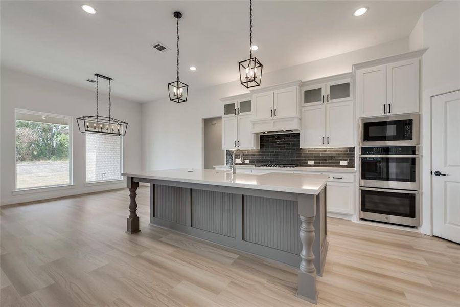 Kitchen with decorative light fixtures, tasteful backsplash, black microwave, an island with sink, and light wood-type flooring