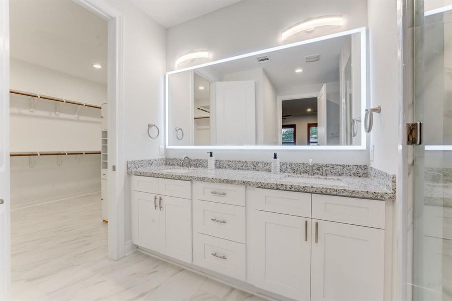 This is a modern and clean bathroom featuring a dual-sink vanity with ample storage, a large mirror with integrated lighting, and a spacious walk-in closet with built-in shelving. The room is finished with sleek white cabinetry and light-toned flooring, giving it a bright and airy feel.