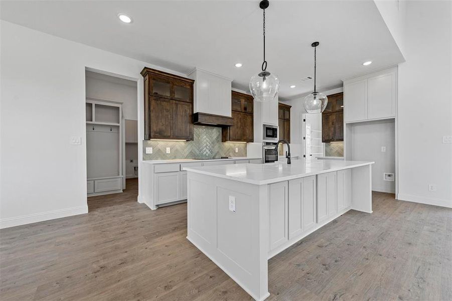 Kitchen featuring an island with sink, stainless steel appliances, light wood-type flooring, backsplash, and hanging light fixtures