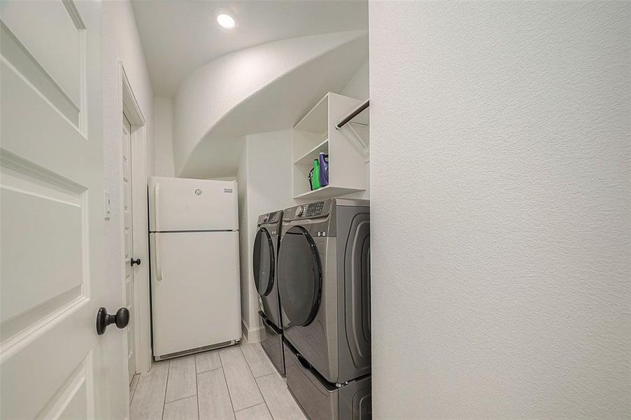 This is a well-kept, modern laundry room featuring a side-by-side washer and dryer set, with a full-size refrigerator and built-in shelving above the appliances. The room is finished with light-colored tile flooring and bright white walls, giving it a clean and airy feel.