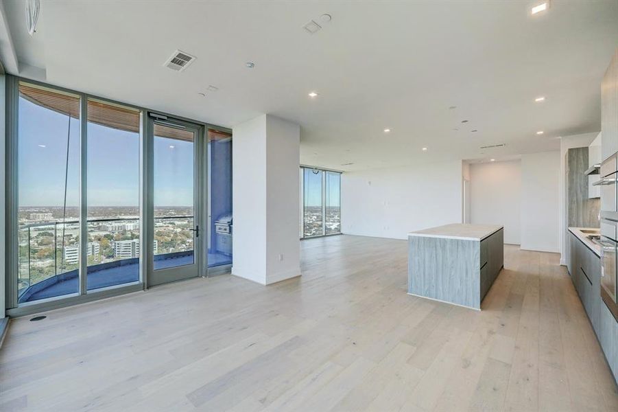 This is a modern, bright  living and dining room with large floor-to-ceiling windows offering expansive city views, complemented by light hardwood floors and a clean, white interior.