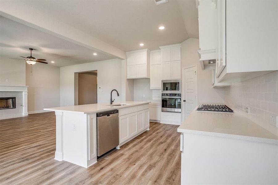 Kitchen featuring appliances with stainless steel finishes, light hardwood / wood-style floors, decorative backsplash, a tiled fireplace, and a kitchen island with sink