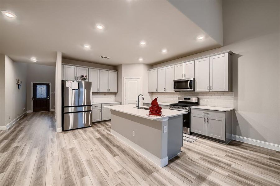 Kitchen with appliances with stainless steel finishes, light hardwood / wood-style floors, tasteful backsplash, and a center island with sink