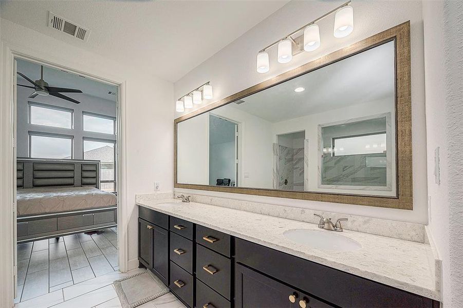 This is a modern bathroom with a large vanity featuring black cabinets and a white countertop, a wide mirror with overhead lighting, and a view into the bedroom with a ceiling fan. The space is bright and neutral-toned.