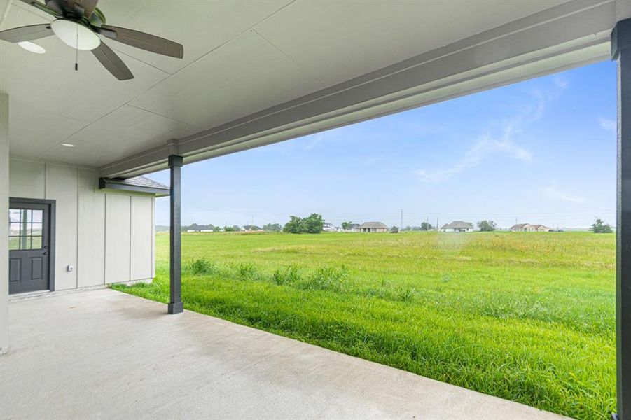 Oversized covered patio space with a ceiling fan, overlooking a spacious and open grassy backyard. There's a view of distant houses and open skies, suggesting a serene, suburban setting.