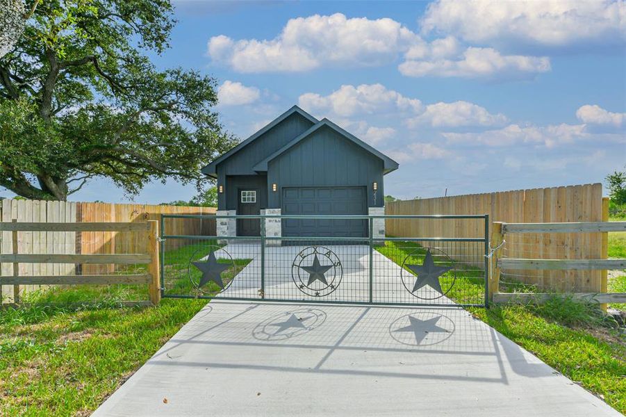 This is a modern single-story home in Beasly Texas. It features a secure gated entry with decorative star elements, a concrete driveway, and a wooden privacy fence surrounding the property. A mature tree adds to the curb appeal.