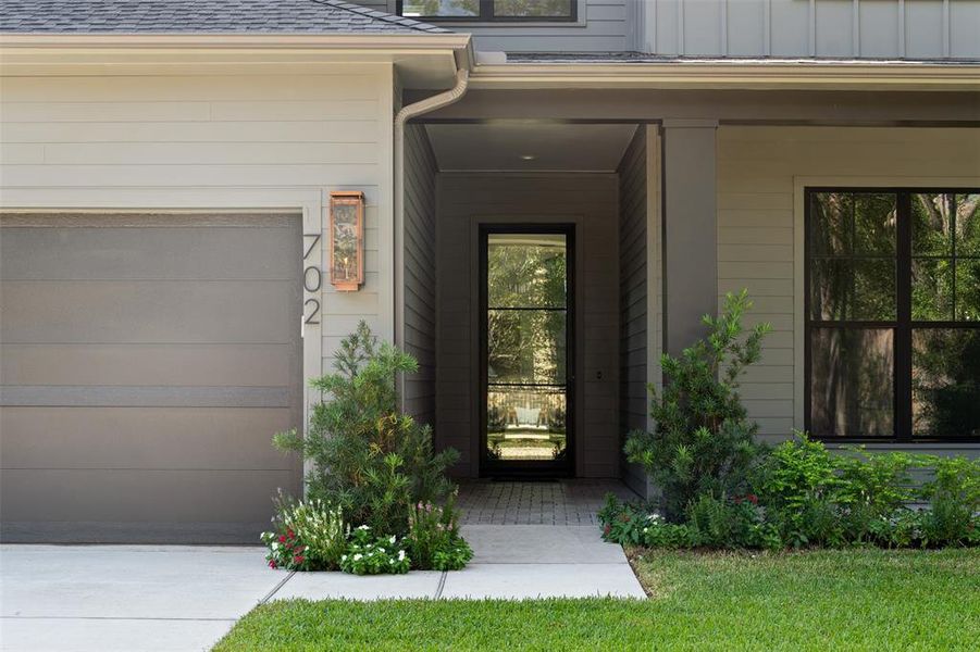 This home features a modern exterior with a neutral color palette, a large garage door, and an inviting entryway framed by lush landscaping. The window provides a glimpse of the greenery outside, enhancing the home's connection with nature.