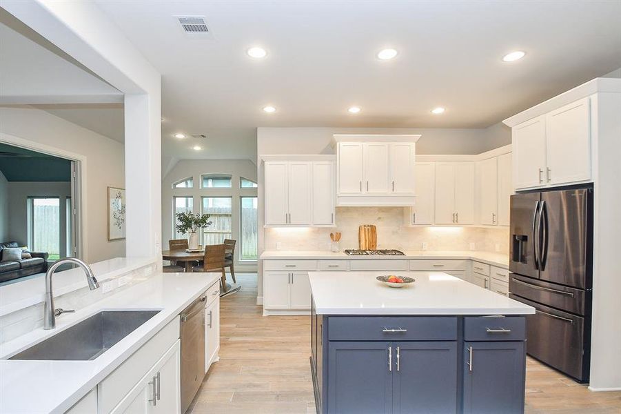 Modern, open-plan kitchen with white cabinetry and a contrasting blue island, featuring stainless steel appliances, recessed lighting, and tile floors. The kitchen opens up to a dining area with ample natural light as well as a large living area.