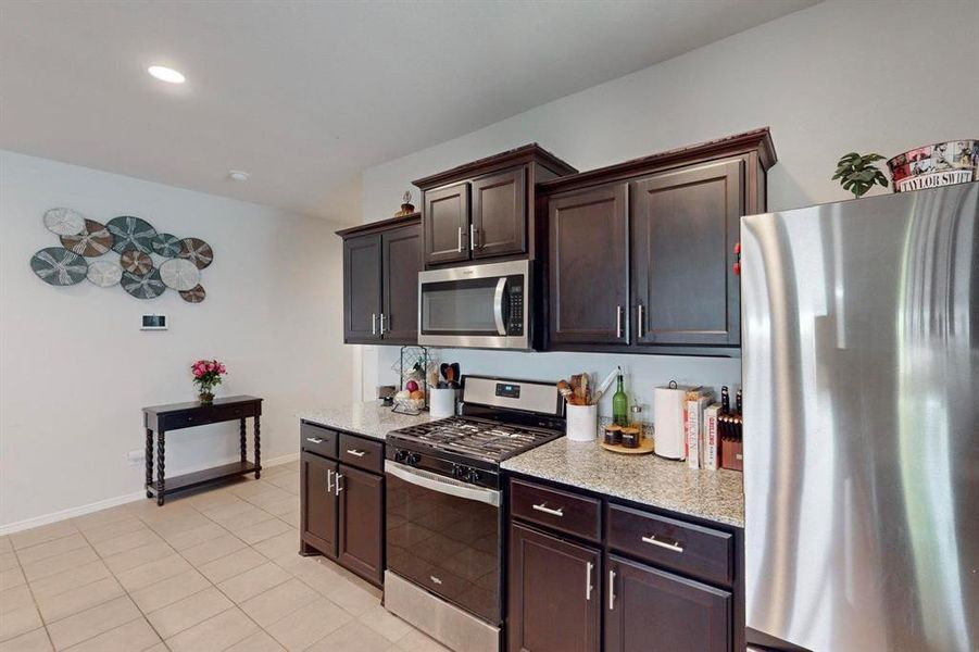 Kitchen featuring light stone countertops, stainless steel appliances, dark brown cabinets, and light tile floors