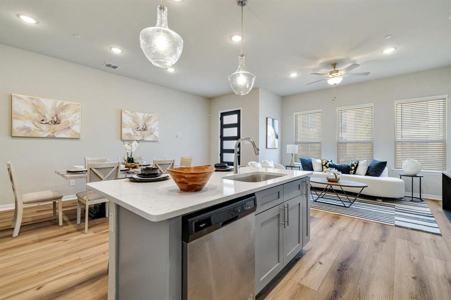 Kitchen with light wood-type flooring, a kitchen island with sink, sink, decorative light fixtures, and stainless steel dishwasher