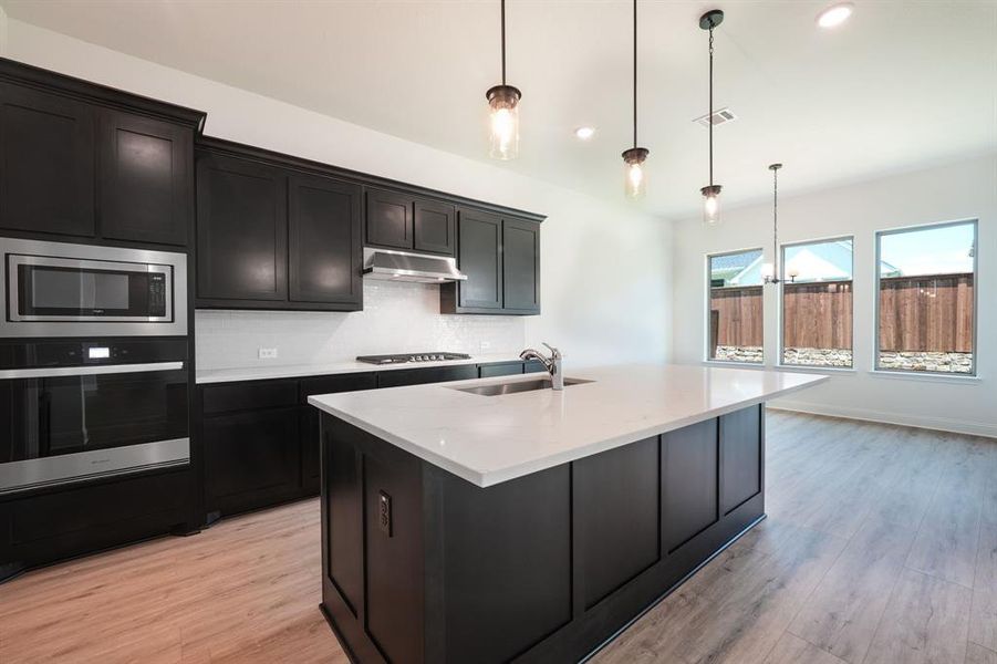 Kitchen featuring decorative light fixtures, stainless steel appliances, backsplash, a kitchen island with sink, and light wood-type flooring