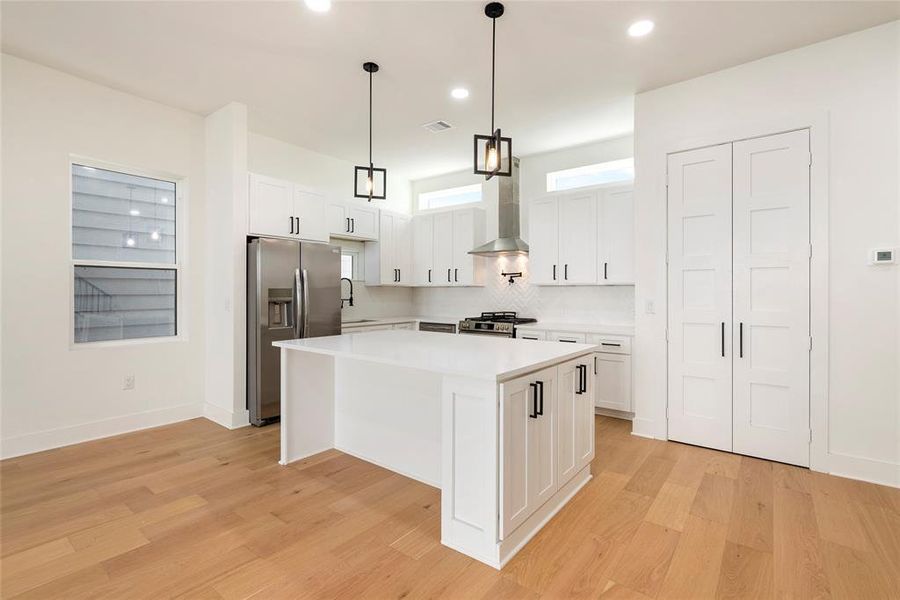 This is a modern, bright kitchen featuring white cabinetry, stainless steel appliances, an island with a sink, stylish pendant lights, and hardwood flooring. There's also a pantry with double doors.