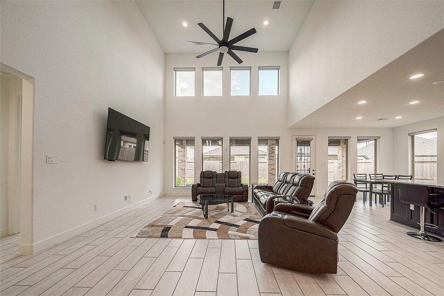 This spacious living room features high ceilings with large windows for ample natural light, a modern ceiling fan, and wood-like tile flooring. It offers a comfortable seating area with leather recliners and is open to the dining space, perfect for entertaining.