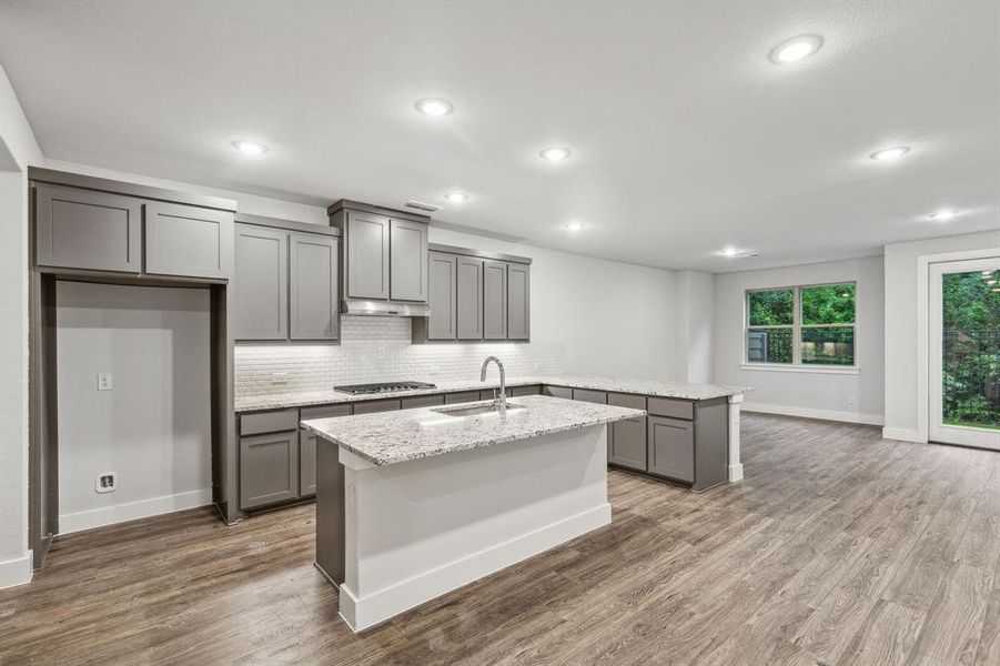Kitchen featuring sink, decorative backsplash, light stone countertops, an island with sink, and wood-type flooring