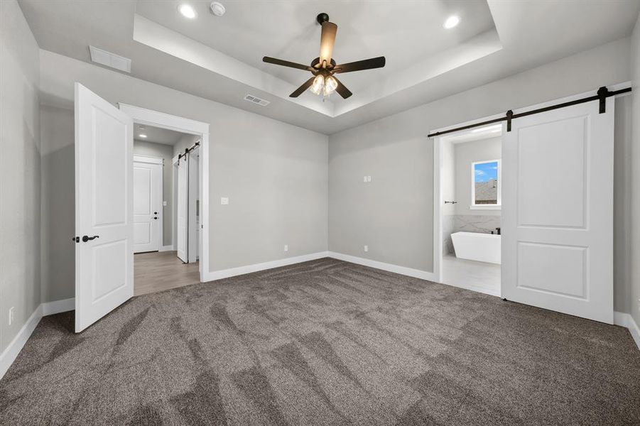 Primary bedroom with ceiling fan, a barn door, dark colored carpet, and a tray ceiling