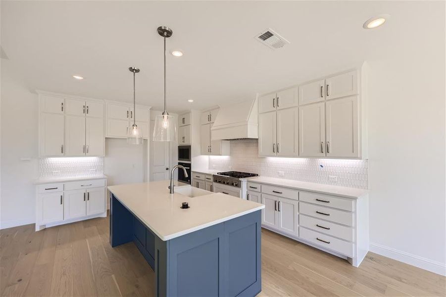 Kitchen featuring custom range hood, light hardwood / wood-style floors, backsplash, a center island with sink, and white cabinetry