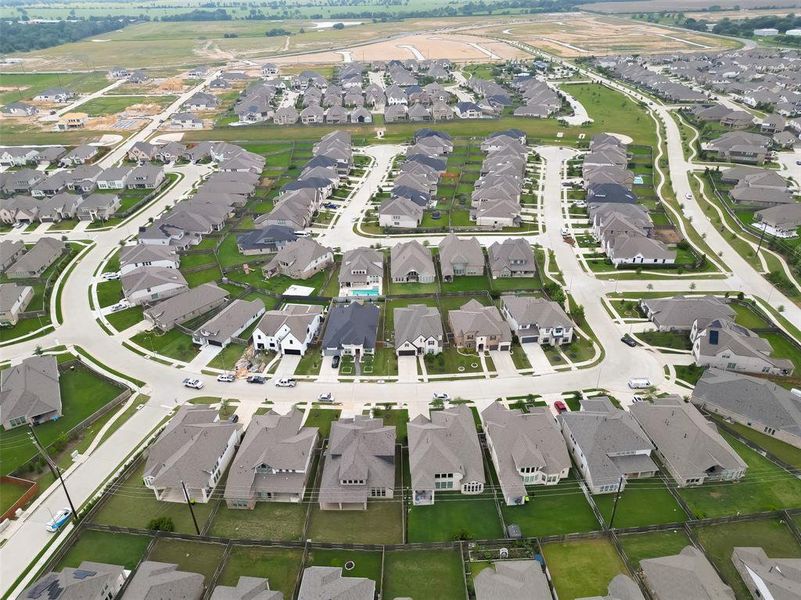 This aerial photo shows a modern suburban neighborhood with numerous detached single-family homes, most with similar architectural styles and gray rooftops, arranged along curved streets and cul-de-sacs. The properties feature well-manicured lawns and driveways, with some homes having backyard fences. The community appears to be recently developed, with additional construction in the background, suggesting potential for growth in the area.