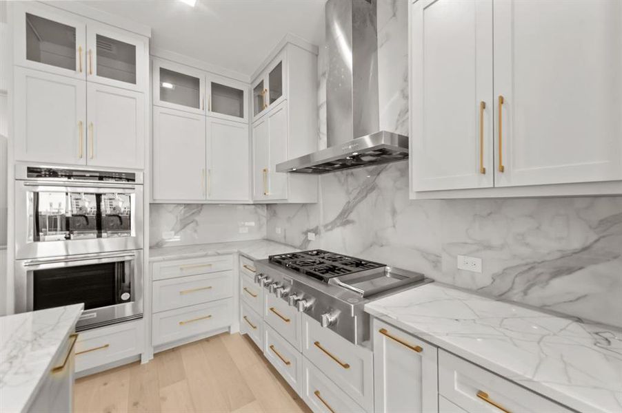 Kitchen with stainless steel appliances, white cabinets, light stone counters, and wall chimney range hood