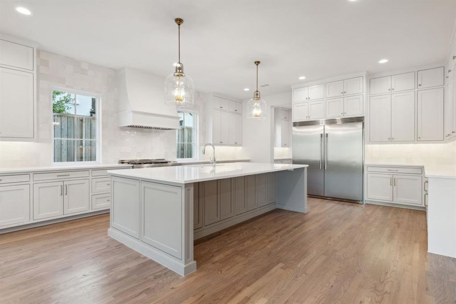 Kitchen with decorative light fixtures, custom exhaust hood, stainless steel built in fridge, an island with sink, and light hardwood / wood-style flooring