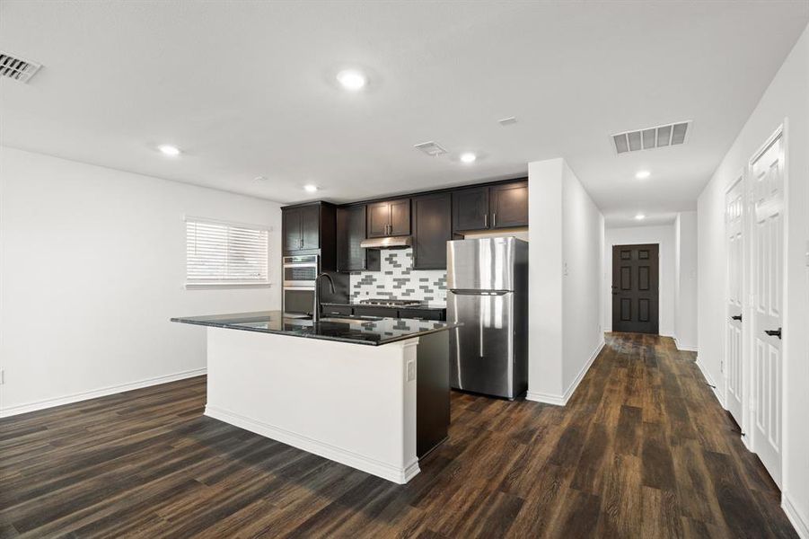 Kitchen featuring dark brown cabinetry, a kitchen island with sink, decorative backsplash, dark wood-type flooring, and appliances with stainless steel finishes