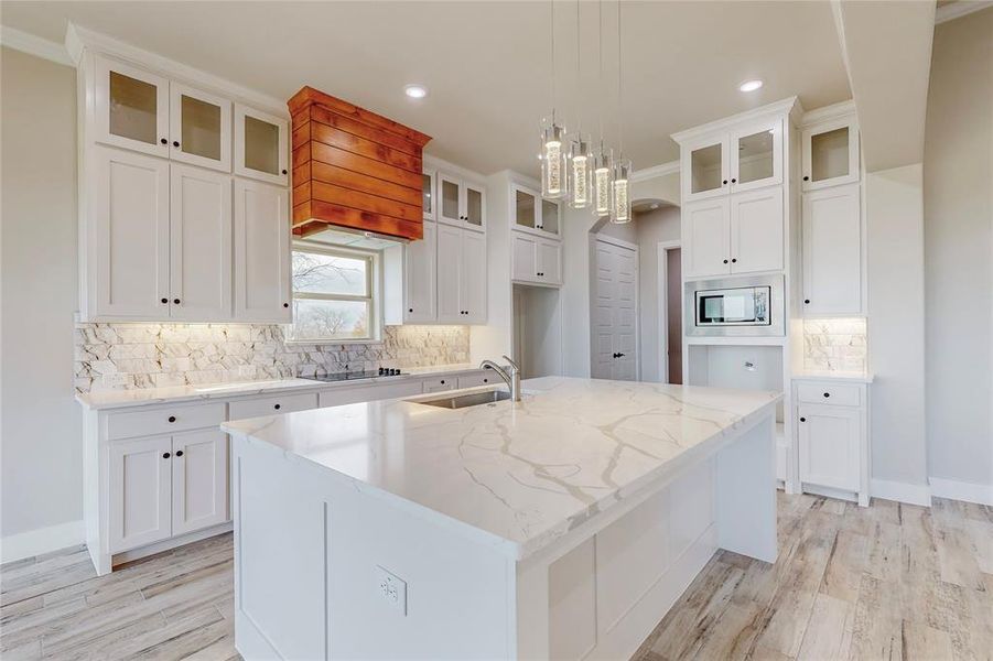 Kitchen featuring stainless steel microwave, light stone counters, white cabinets, crown molding, and a kitchen island with sink