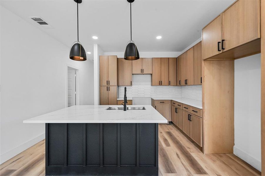 Kitchen featuring light hardwood / wood-style floors, a kitchen island with sink, light stone counters, backsplash, and pendant lighting