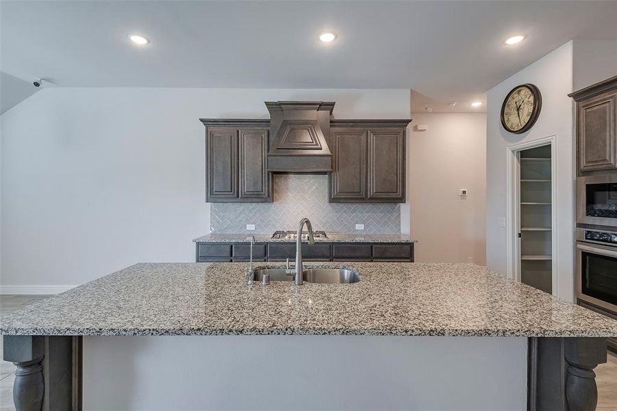 Kitchen with appliances with stainless steel finishes, sink, dark brown cabinets, premium range hood, and a center island with sink