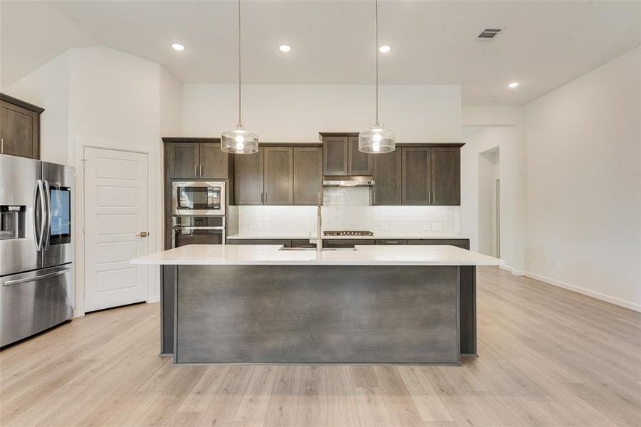 Kitchen featuring stainless steel appliances, an island with sink, dark brown cabinetry, and light hardwood / wood-style flooring