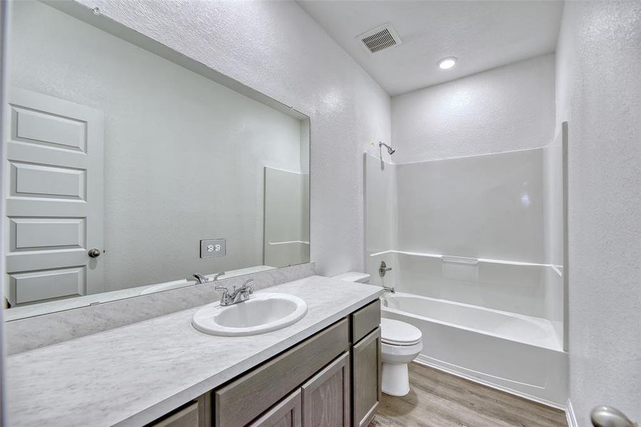 The secondary bathroom offers a spacious counter with ample drawers and cabinets, ideal for shared use. A large frameless mirror hangs above the vanity, enhancing the room's spacious feel and functionality. An oversized tub/shower combination stands ready for relaxation or quick refreshment.