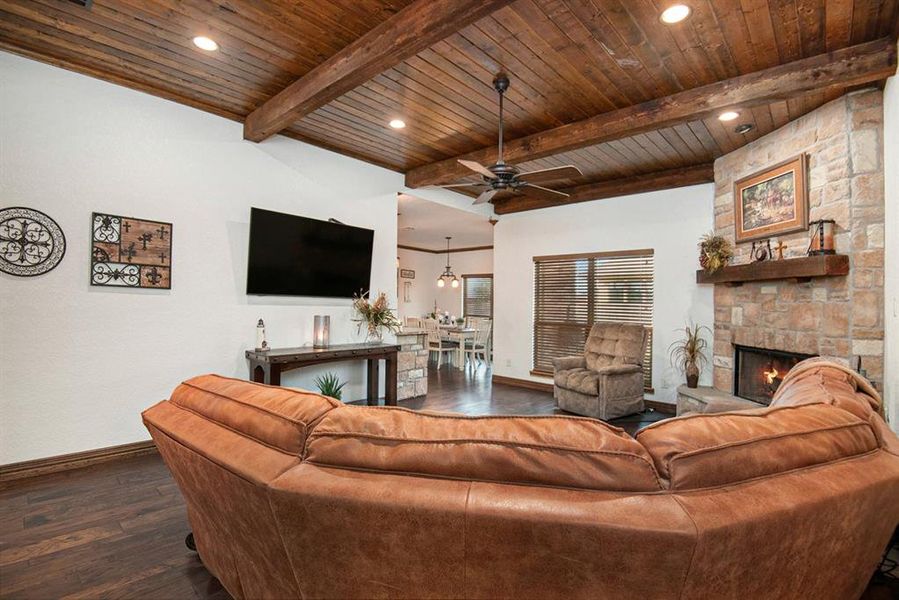 Living room with a stone fireplace, dark hardwood / wood-style flooring, ceiling fan, wooden ceiling, and beamed ceiling