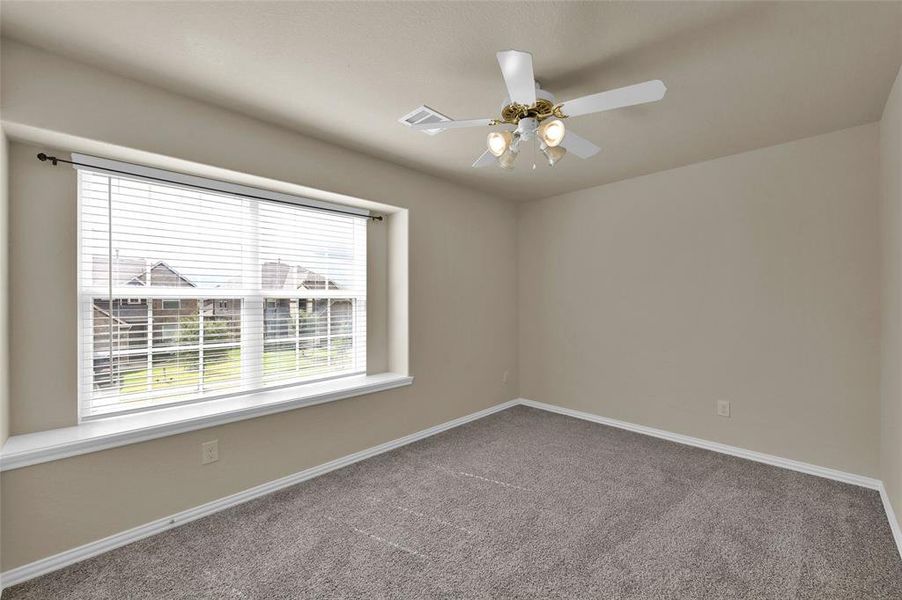 This is a well-lit, empty room featuring a large window with blinds, neutral walls, plush carpeting, and a ceiling fan with lights.